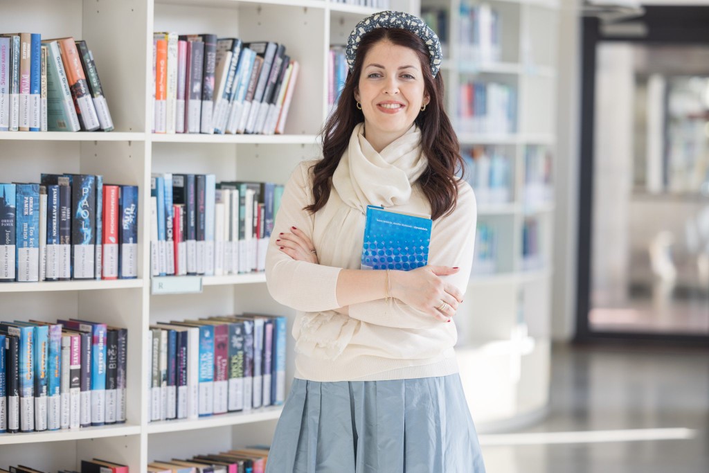 Dr Marijana Prodanović standing in a library, smiling and holding a book